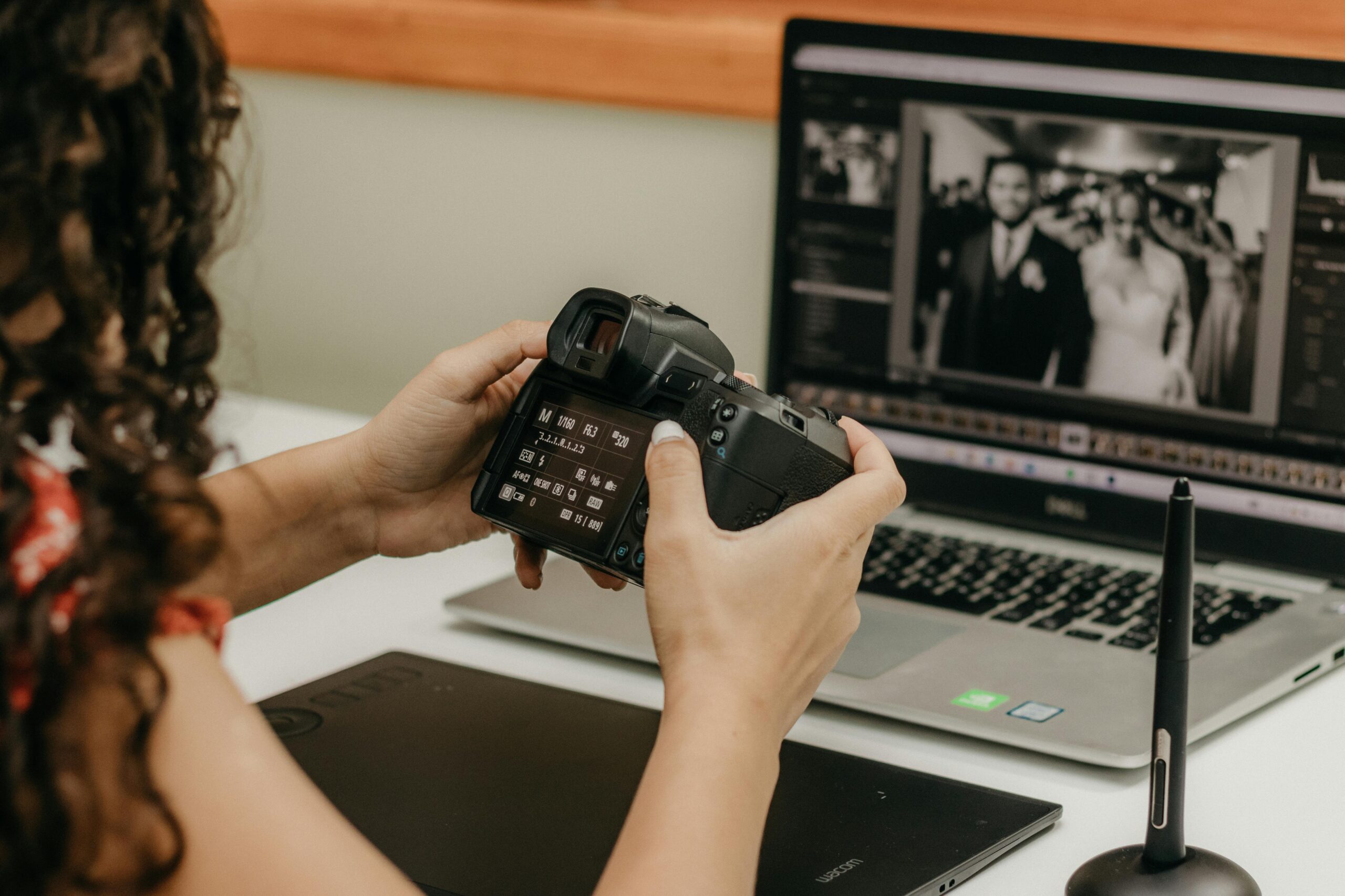 A woman edits wedding photos on a laptop, holding a DSLR camera and using a graphics tablet.