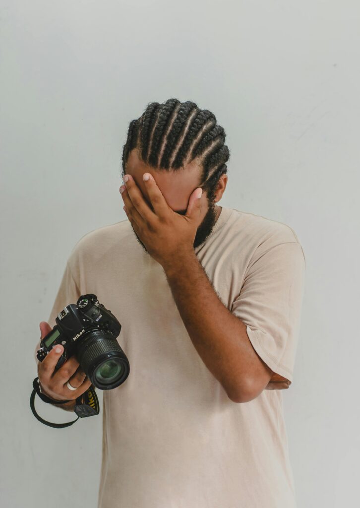 A photographer in a beige shirt facepalming while holding a camera, showing dismay or embarrassment.