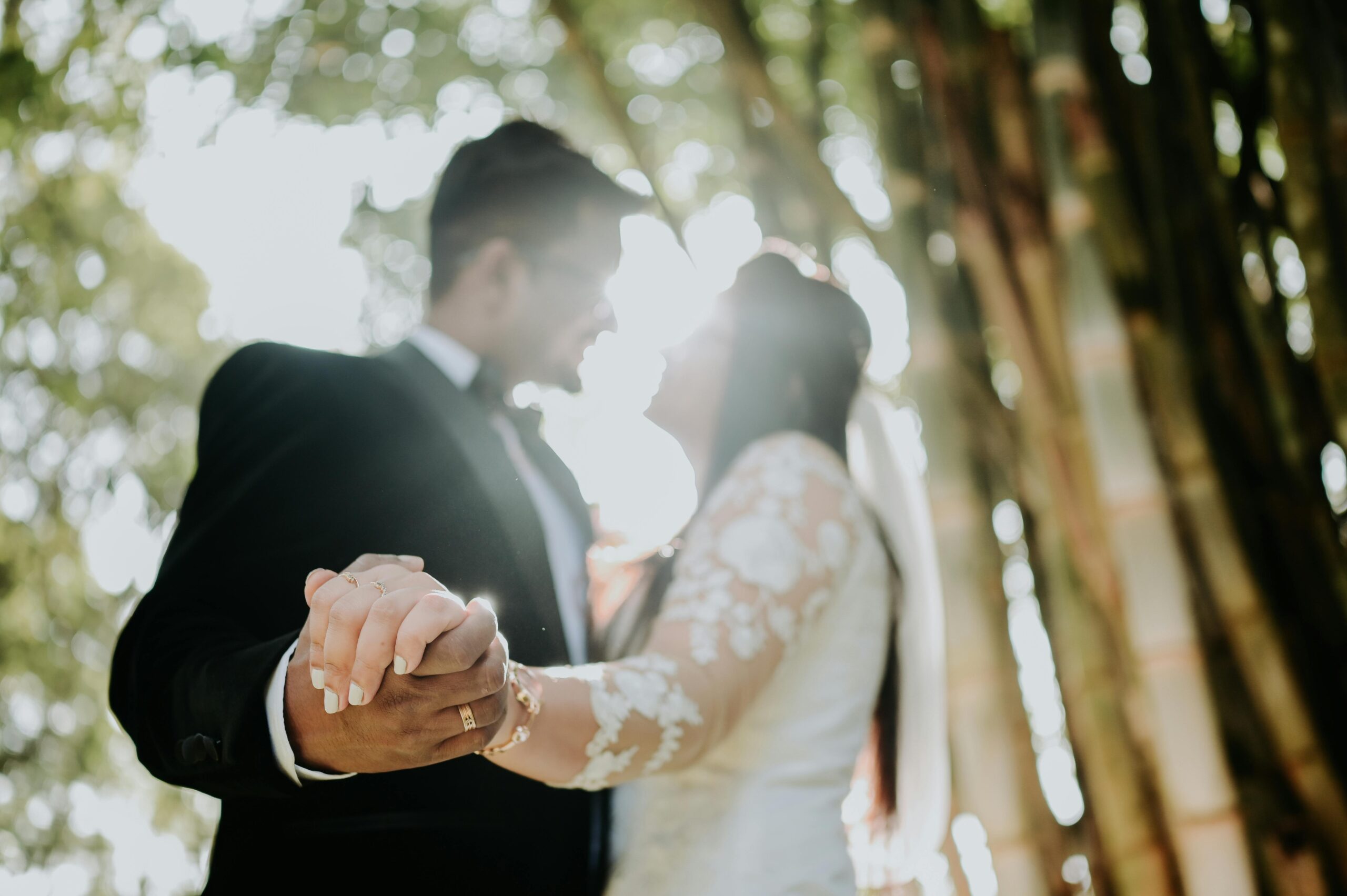 A romantic moment of a newlywed couple holding hands under sunlight among trees.