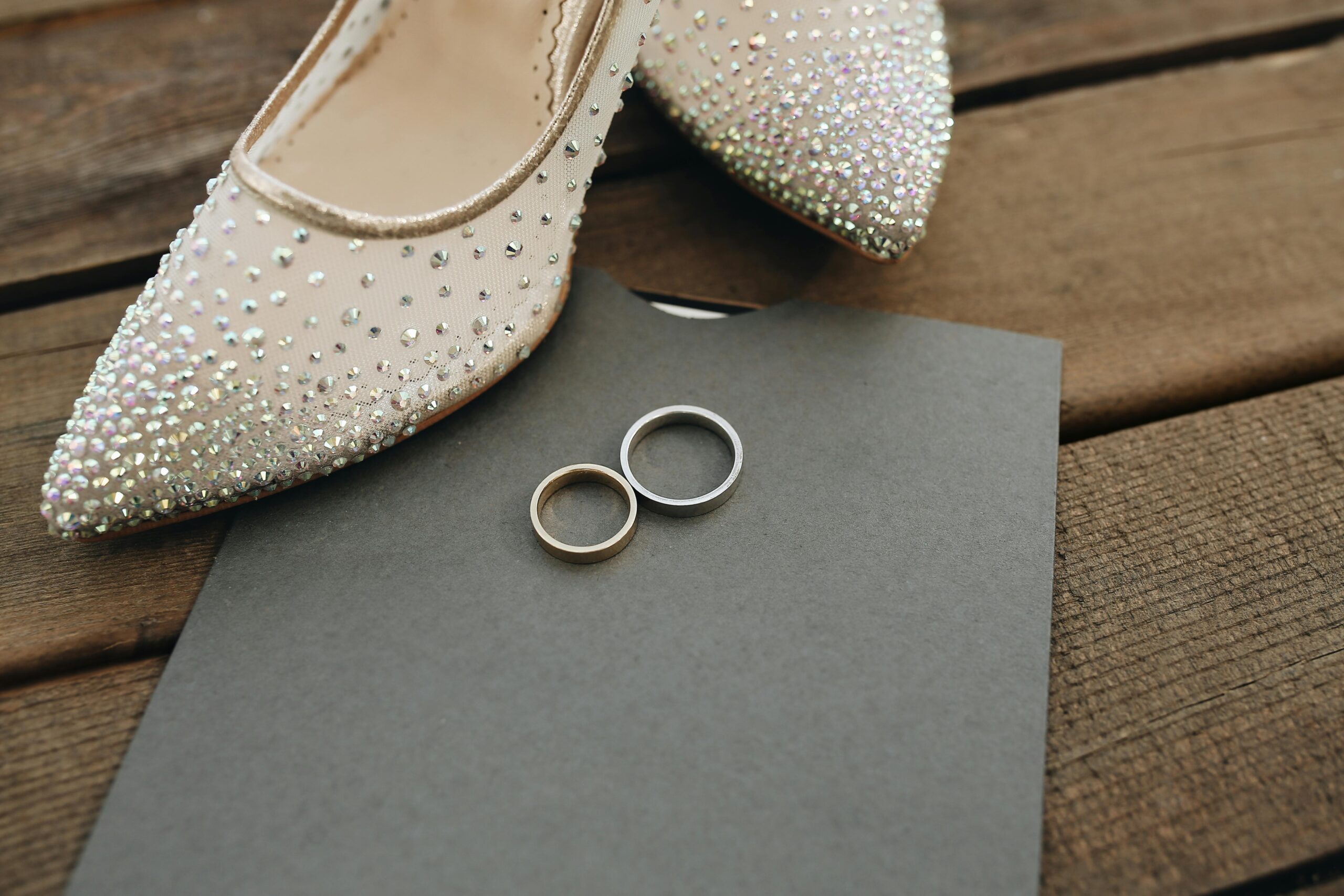 Close-up of sparkling bridal shoes and wedding rings on wood surface.
