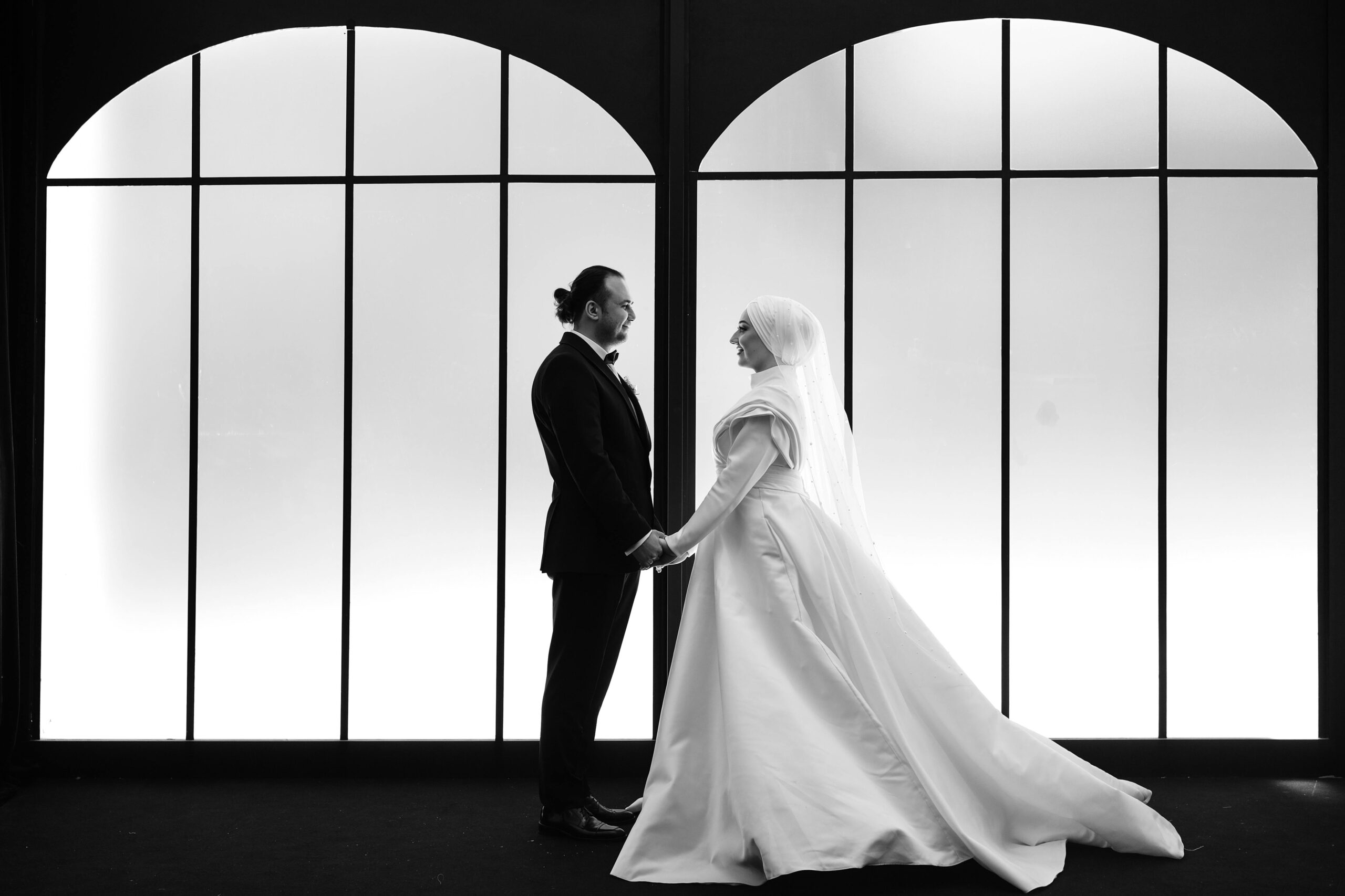 A black and white photo of a couple in wedding attire standing indoors, holding hands.