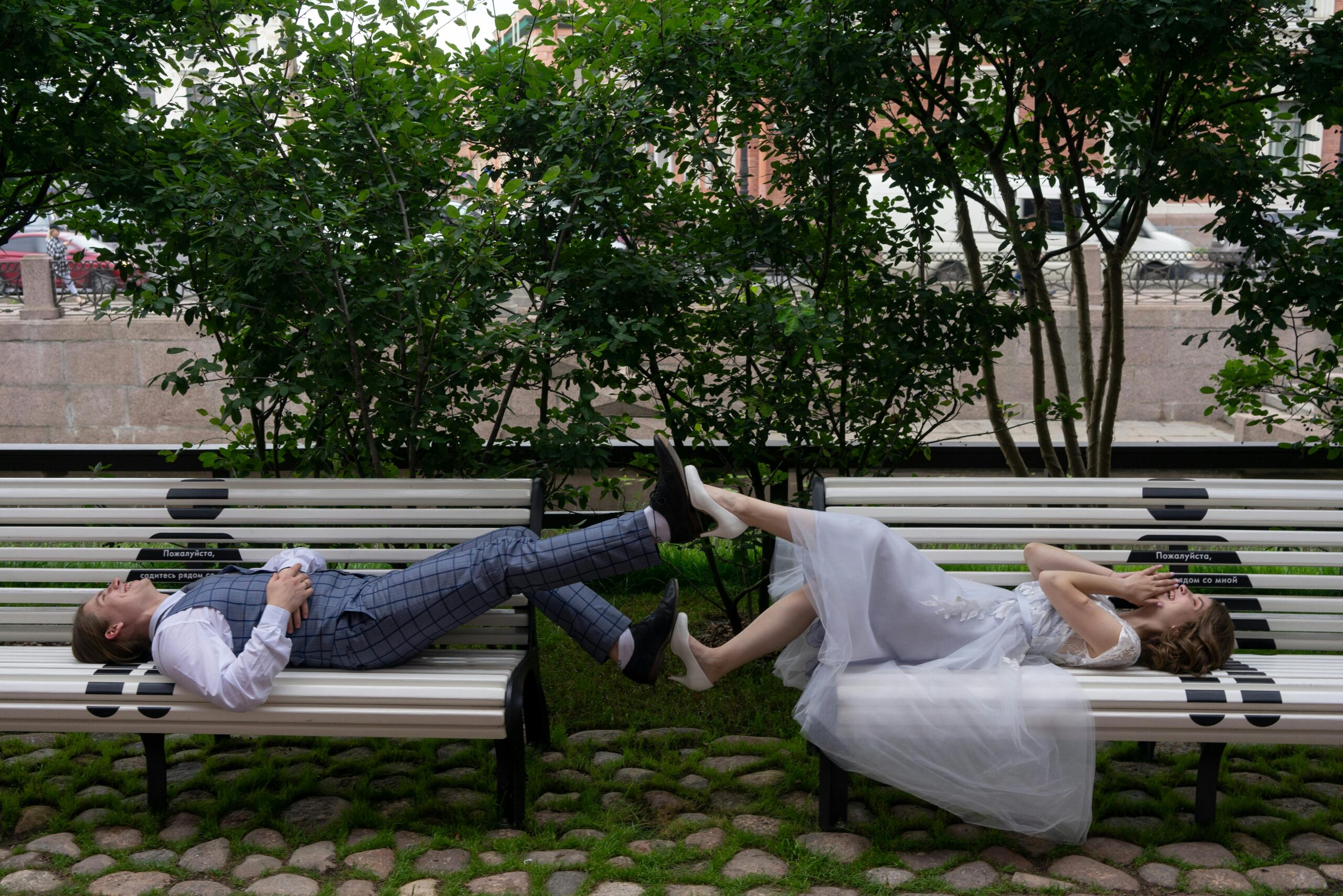 Side view of young cheerful couple in stylish apparel touching legs of each other while resting on urban benches